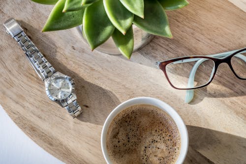 A Cup Of Coffee, Stainless Watch And Eyeglasses On A Table With Ornamental Plant