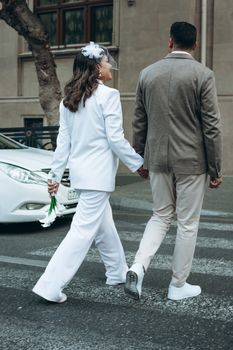 Newlywed couple holding hands and crossing a city street, showcasing modern wedding fashion.