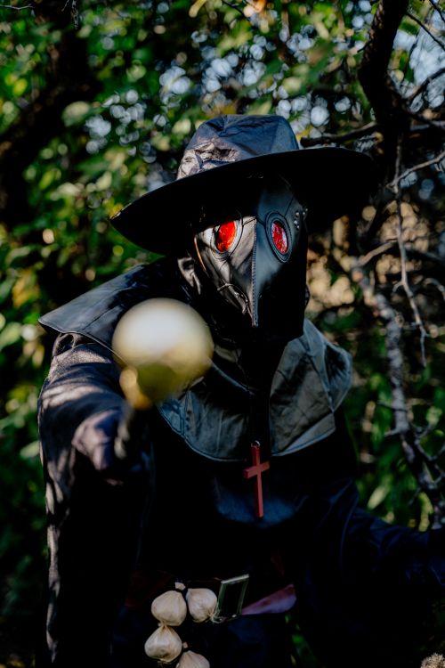 Free A figure in a plague doctor costume holding an object in a shadowy garden. Stock Photo