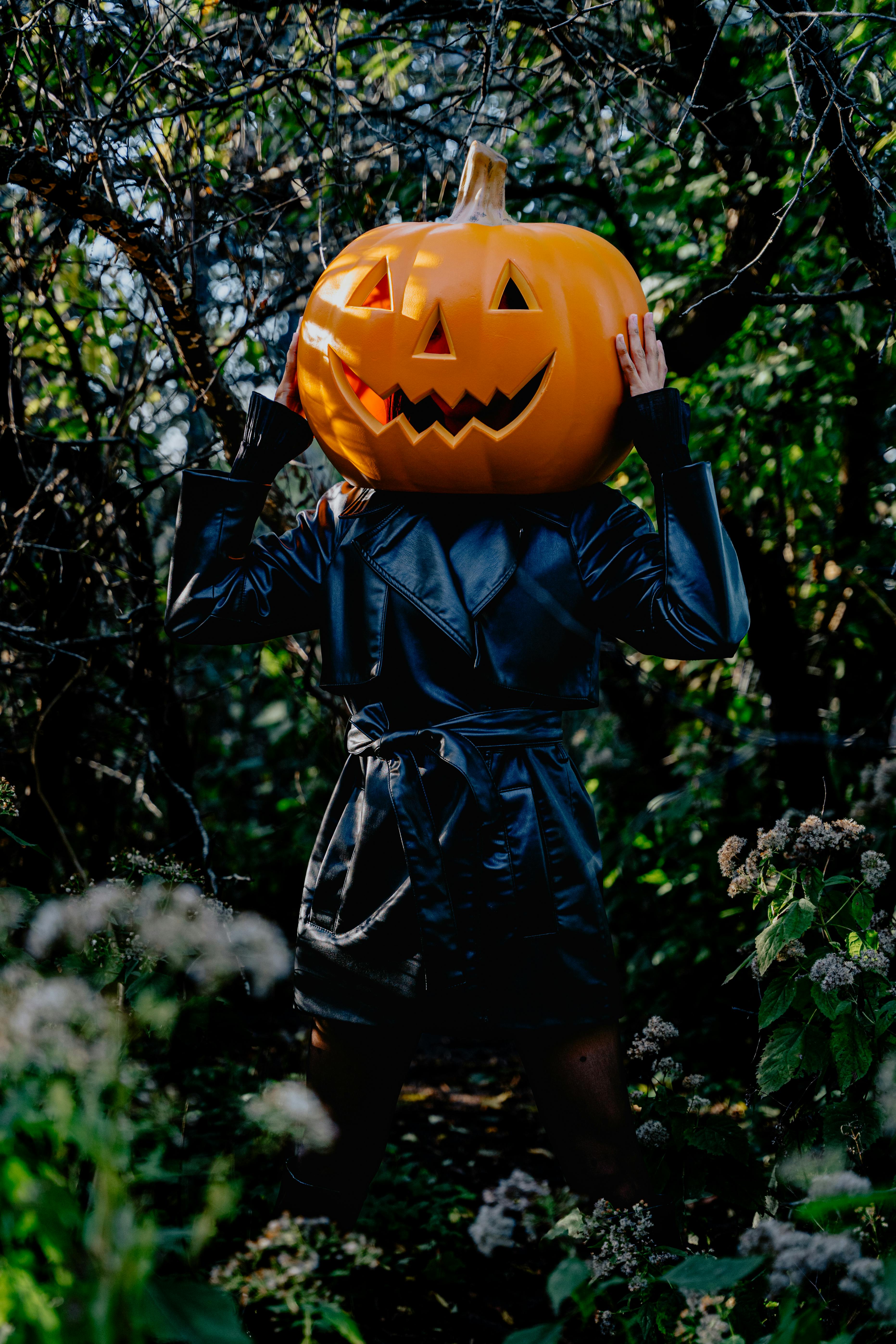 Person wearing a black coat and a pumpkin head in a dark, spooky outdoor setting.