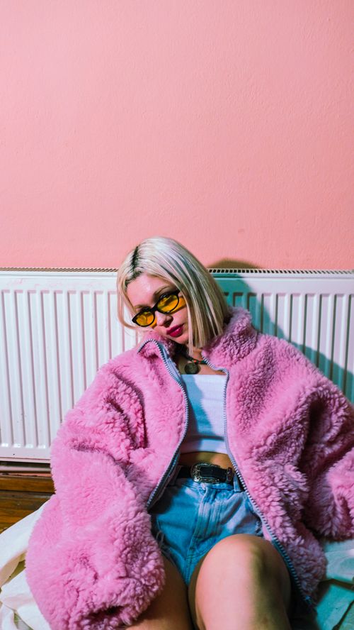 Free Fashionable young woman in a pink fuzzy jacket and sunglasses posing stylishly indoors. Stock Photo