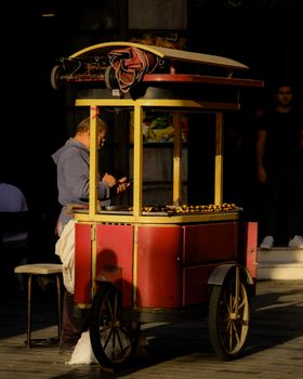 A man tending a vibrant red street food cart selling roasted chestnuts in an urban setting.