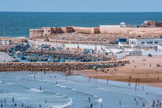 Aerial capture of a lively beach with people enjoying the waves near a historical fortress.