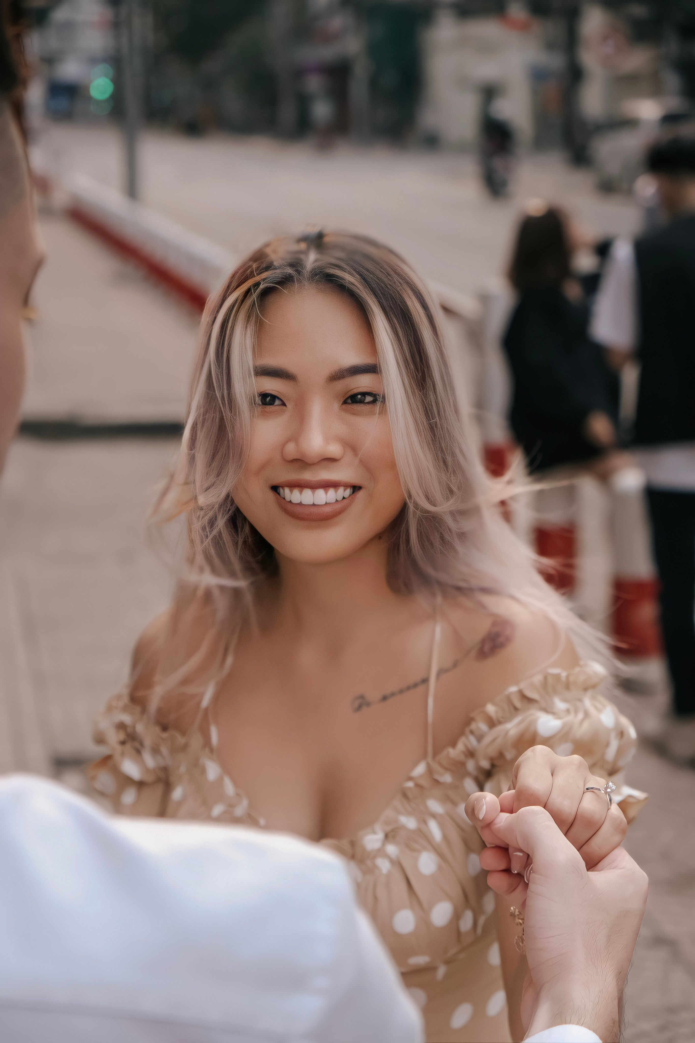A smiling young woman in a polka dot dress is captured holding hands outdoors.