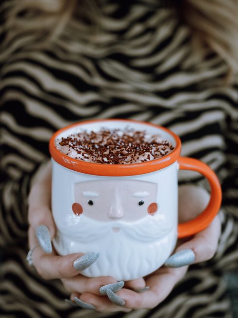 Close-up of a Santa-themed mug with hot chocolate topped with whipped cream and sprinkles, held by a woman.