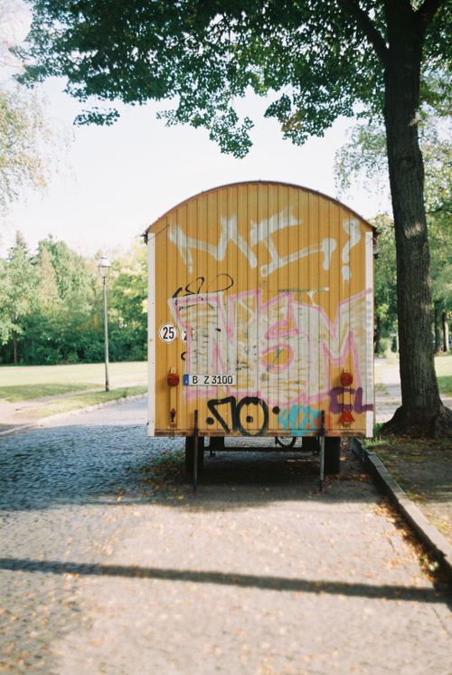 Free Graffitied yellow trailer parked on cobblestone path in a sunlit park, surrounded by trees. Stock Photo
