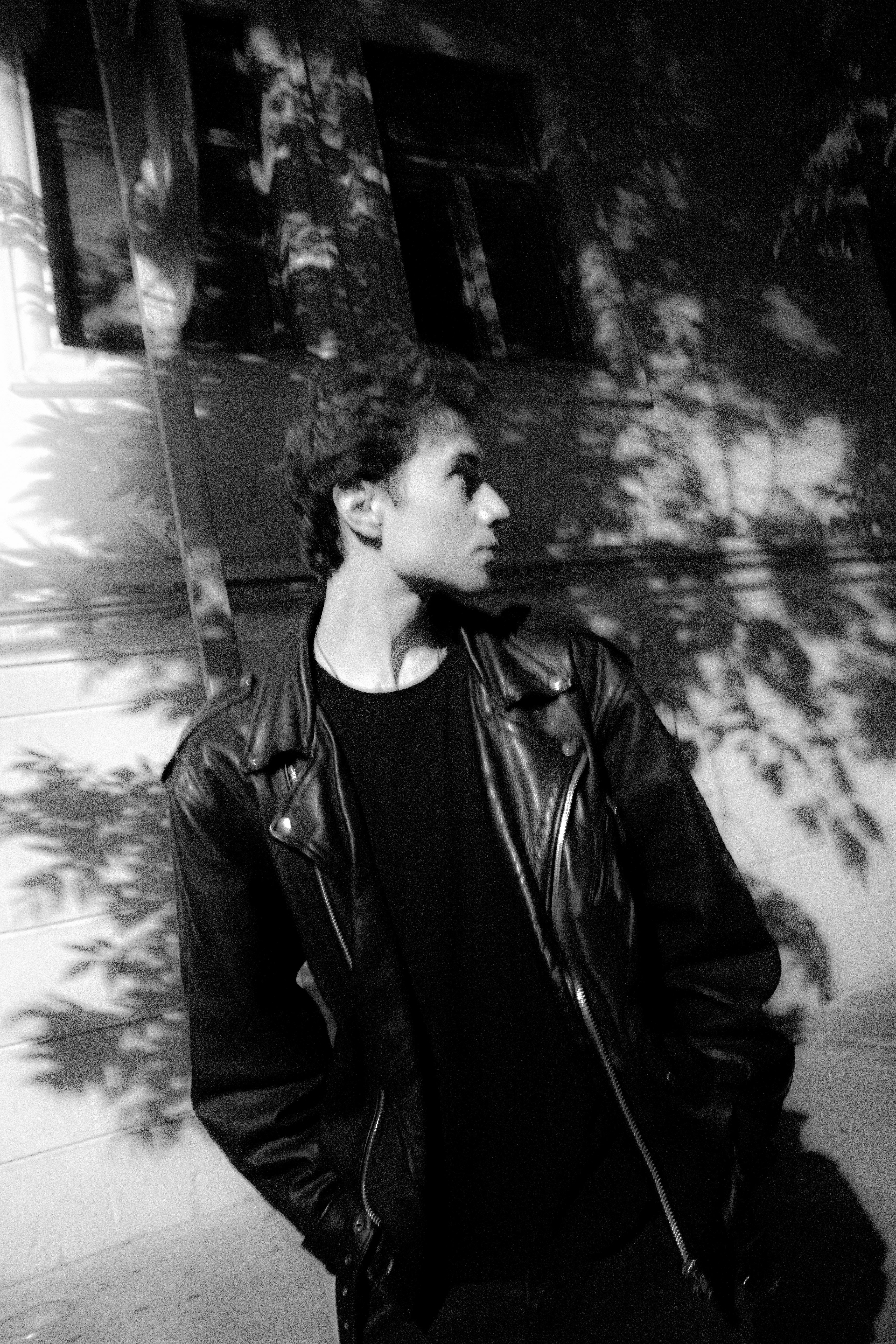 A black and white photo of a man in a leather jacket standing on a city sidewalk at night.