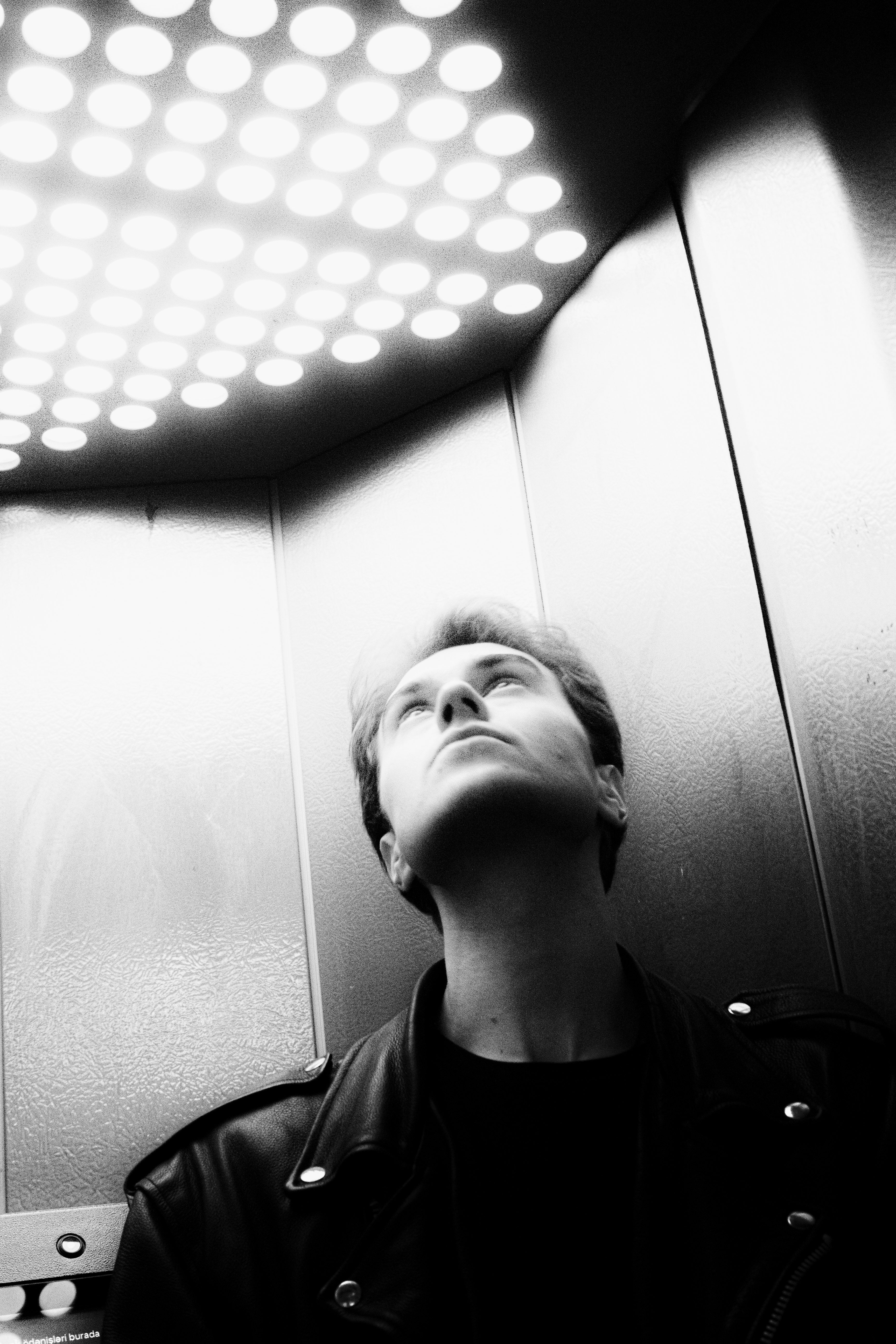 A man in a leather jacket looks up at illuminated lights in an elevator.