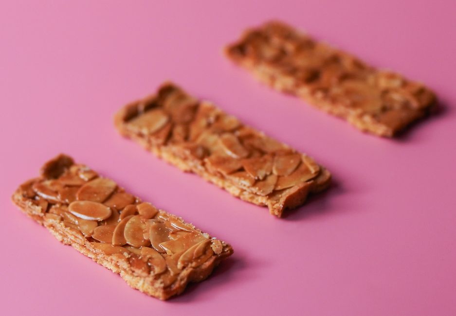 Thin, golden almond tuile cookies draped over a rolling pin so they cool into a curved shape, with lacy caramelized edges visible against the light.