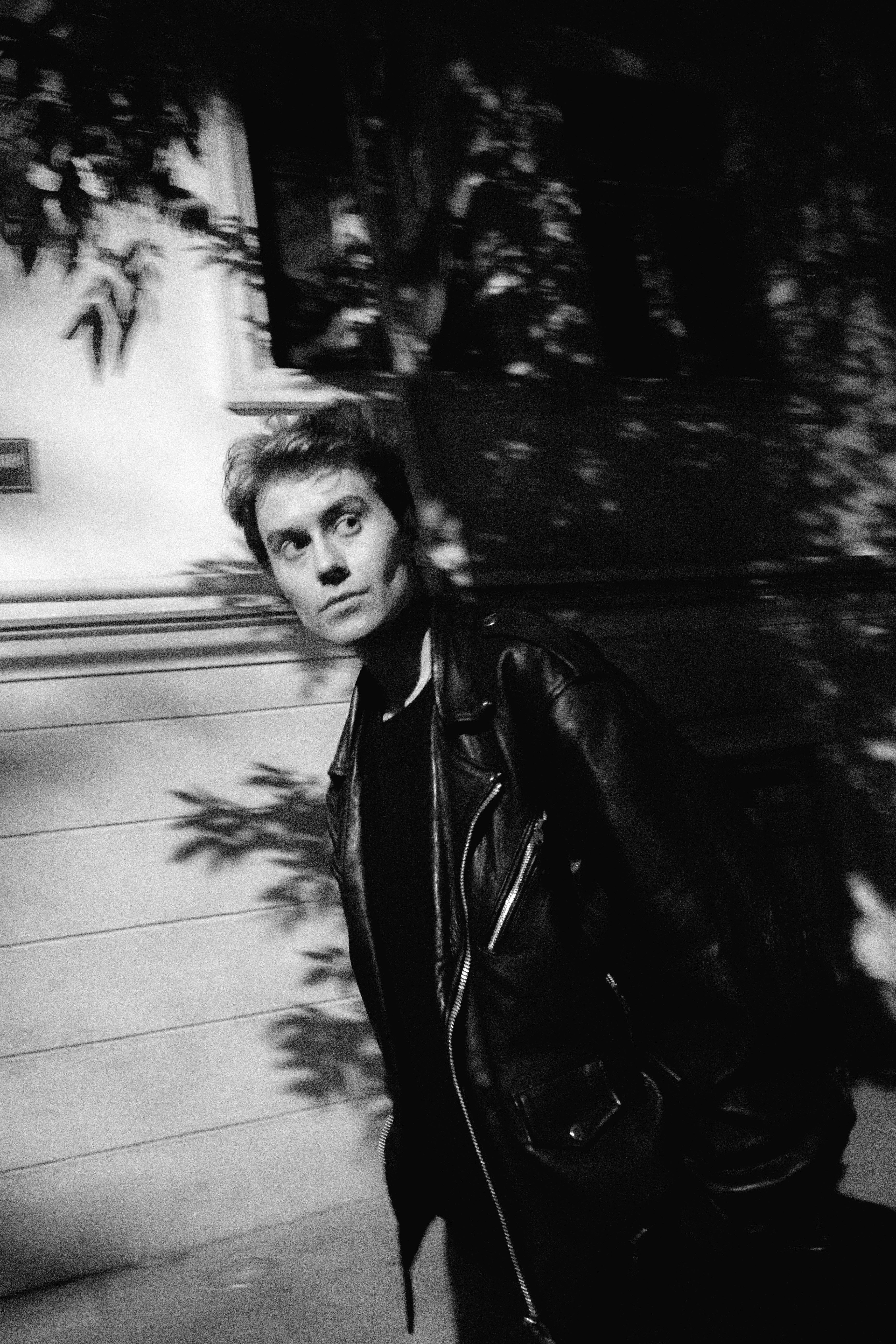 Black and white photo of a stylish man wearing a leather jacket walking outdoors.
