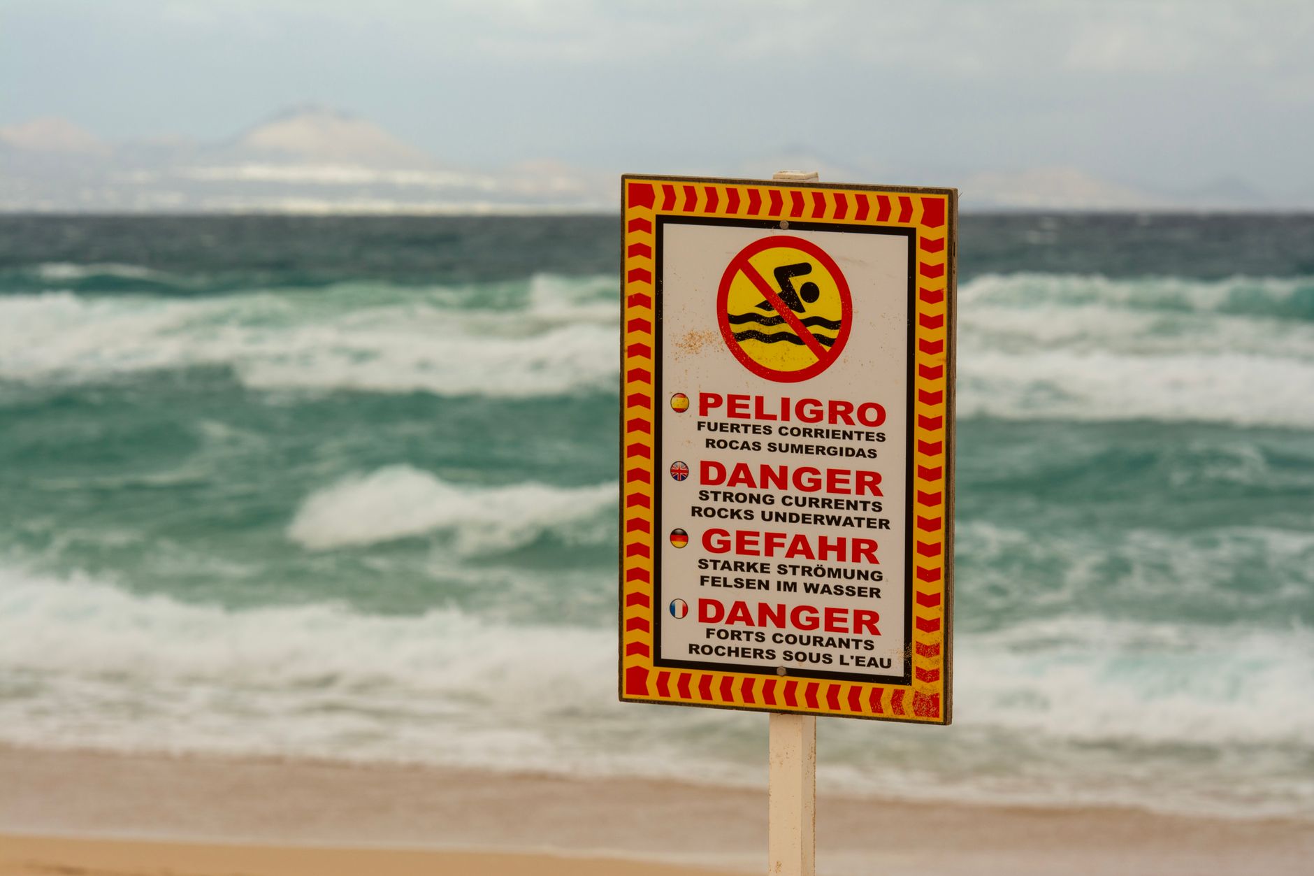 Un cartel de advertencia de playa advirtiendo sobre fuertes corrientes submarinas en múltiples idiomas.