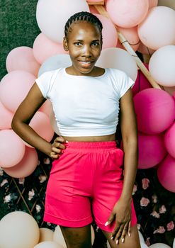 Smiling woman in a white top and red shorts posing with pink balloons, exuding happiness and style.