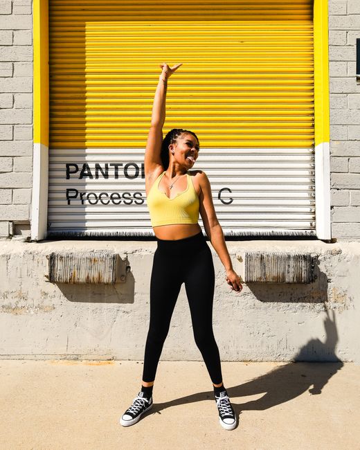 A vibrant young woman in sportswear joyfully posing outside a warehouse on a sunny day.