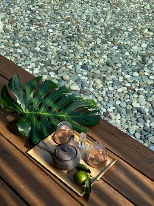 Free A peaceful setting with a tea set on a tray beside a Monstera leaf, overlooking a pebble-covered pool. Stock Photo