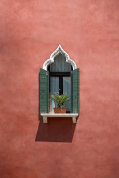 A charming Venetian window with green shutters and a potted plant on a red stucco wall.
