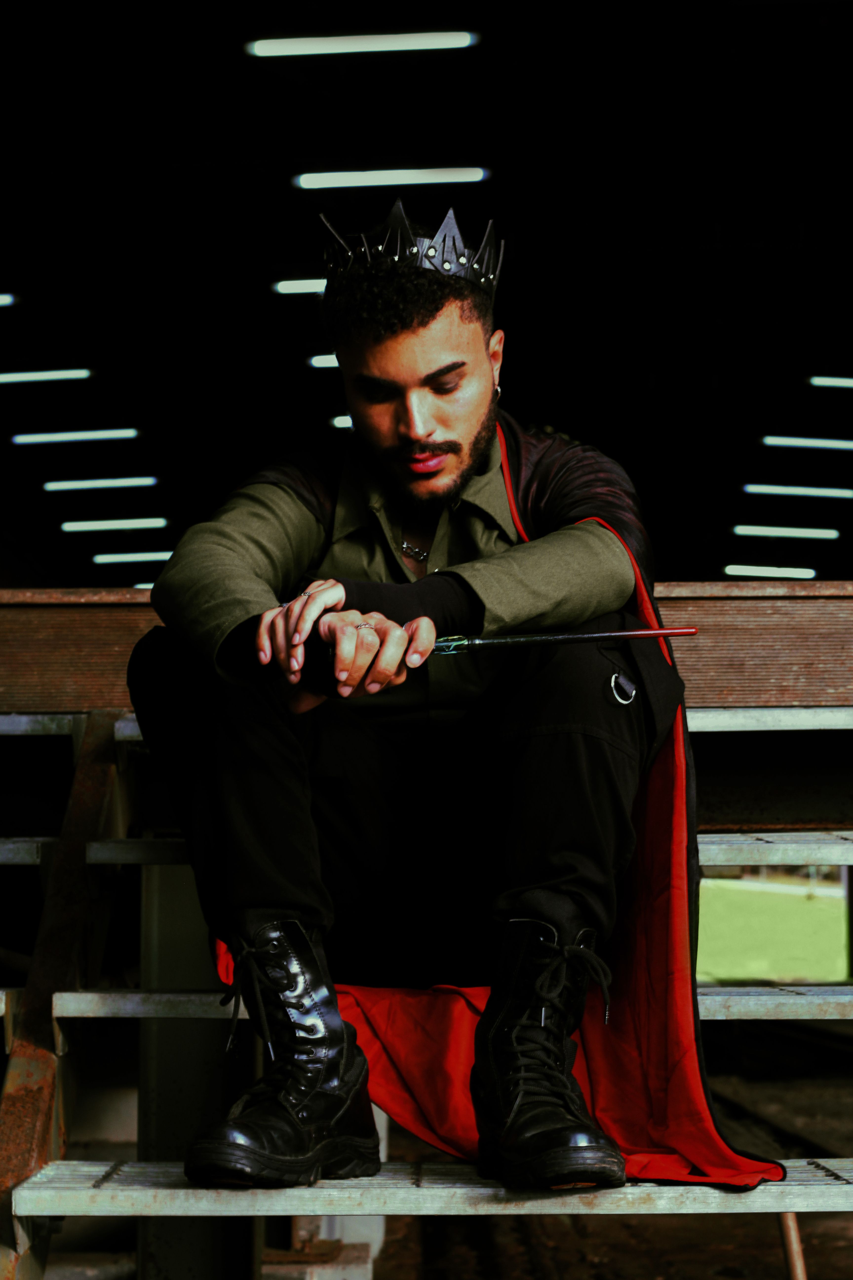 A dramatic portrait featuring a man in fantasy attire with a crown and wand, sitting pensively indoors.