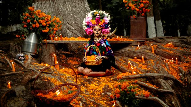 A traditional Dia de los Muertos celebration with a woman in colorful attire sitting outdoors surrounded by marigolds and candles.