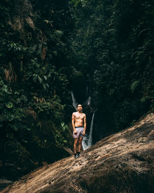 Free A topless man stands on a rock in a dense forest with a waterfall backdrop, embracing nature. Stock Photo