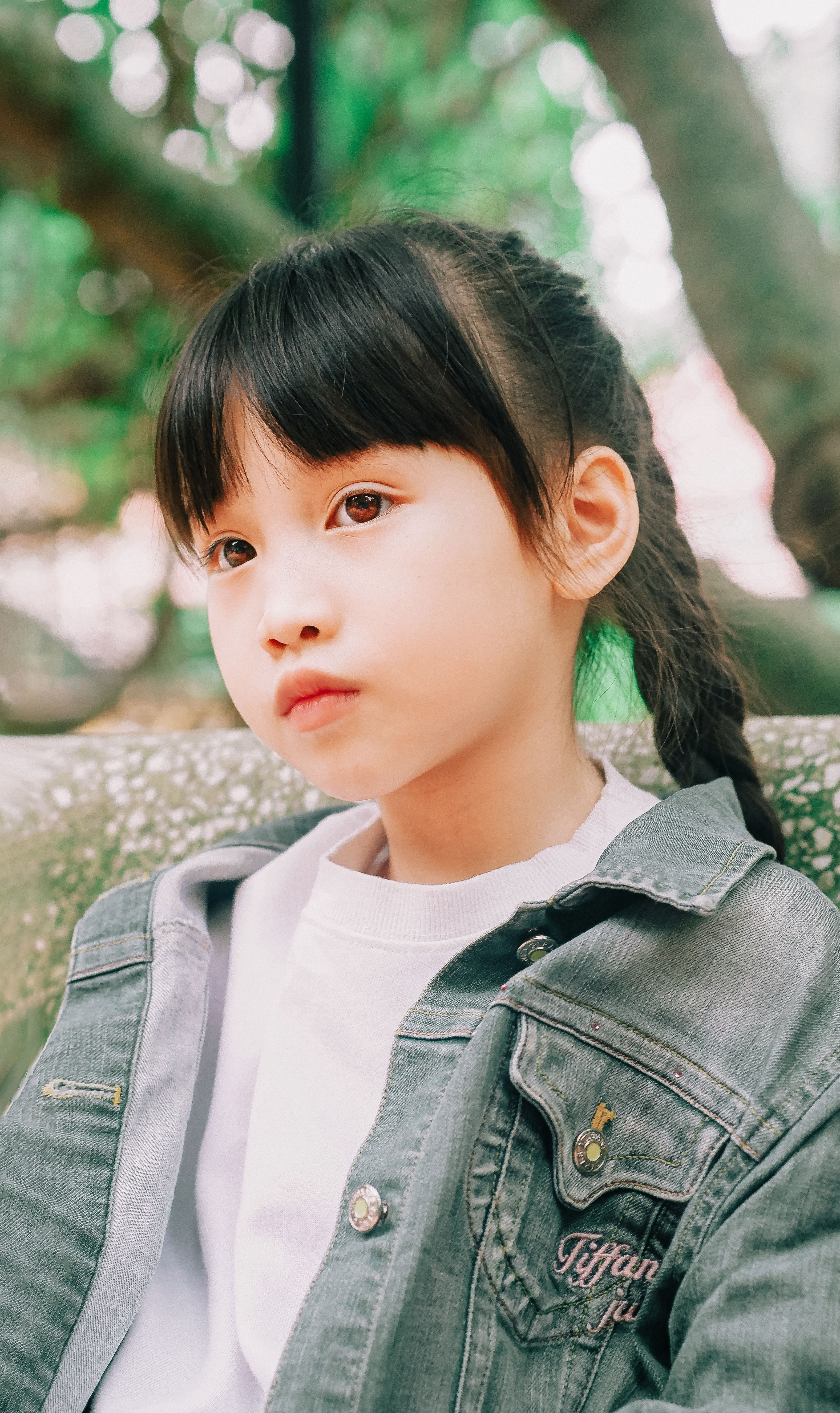 Charming portrait of a young girl with braided hair wearing a denim jacket, posing outdoors in a green park.