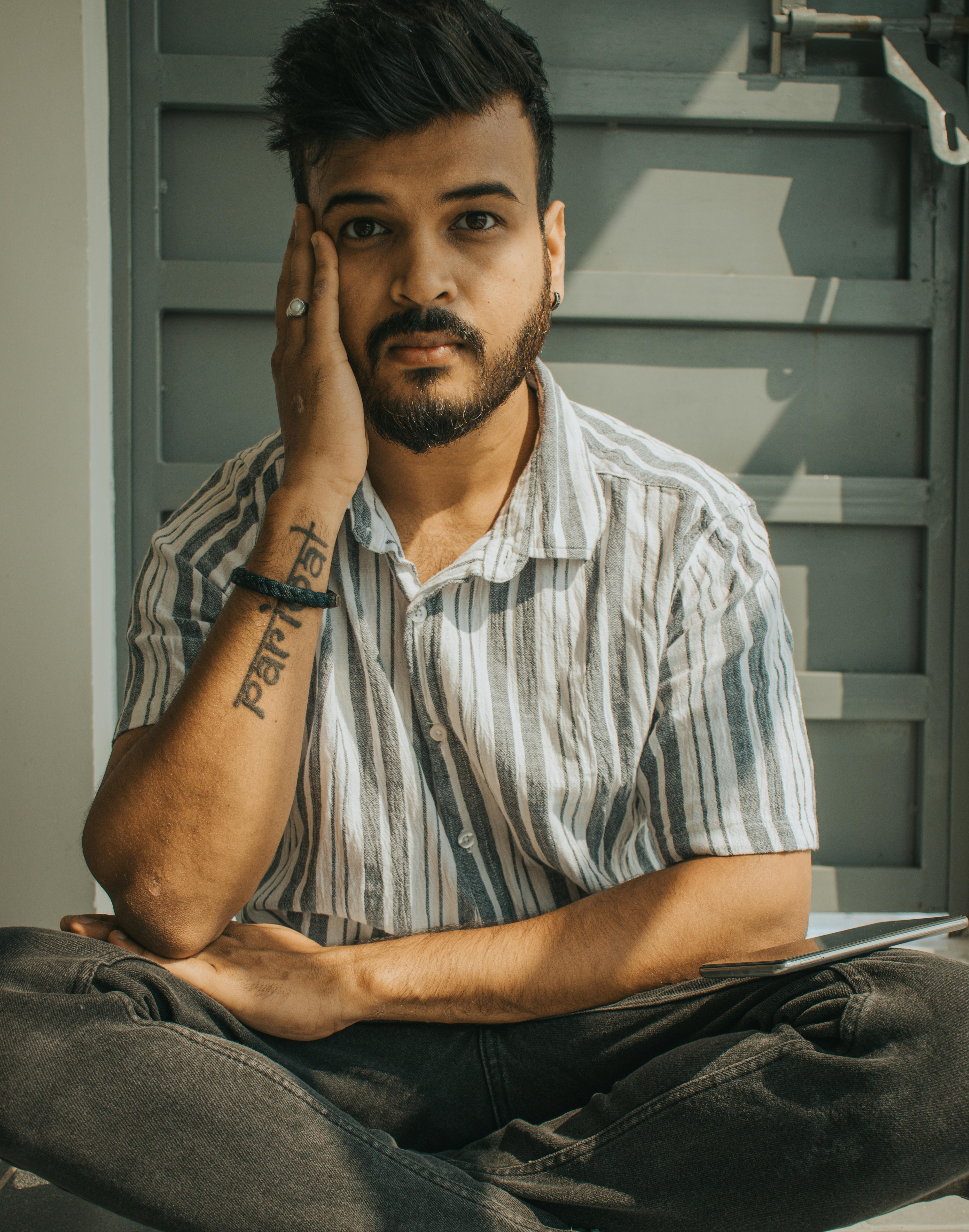 Casual portrait of a bearded man sitting cross-legged, showcasing a stylish striped shirt and tattoo.