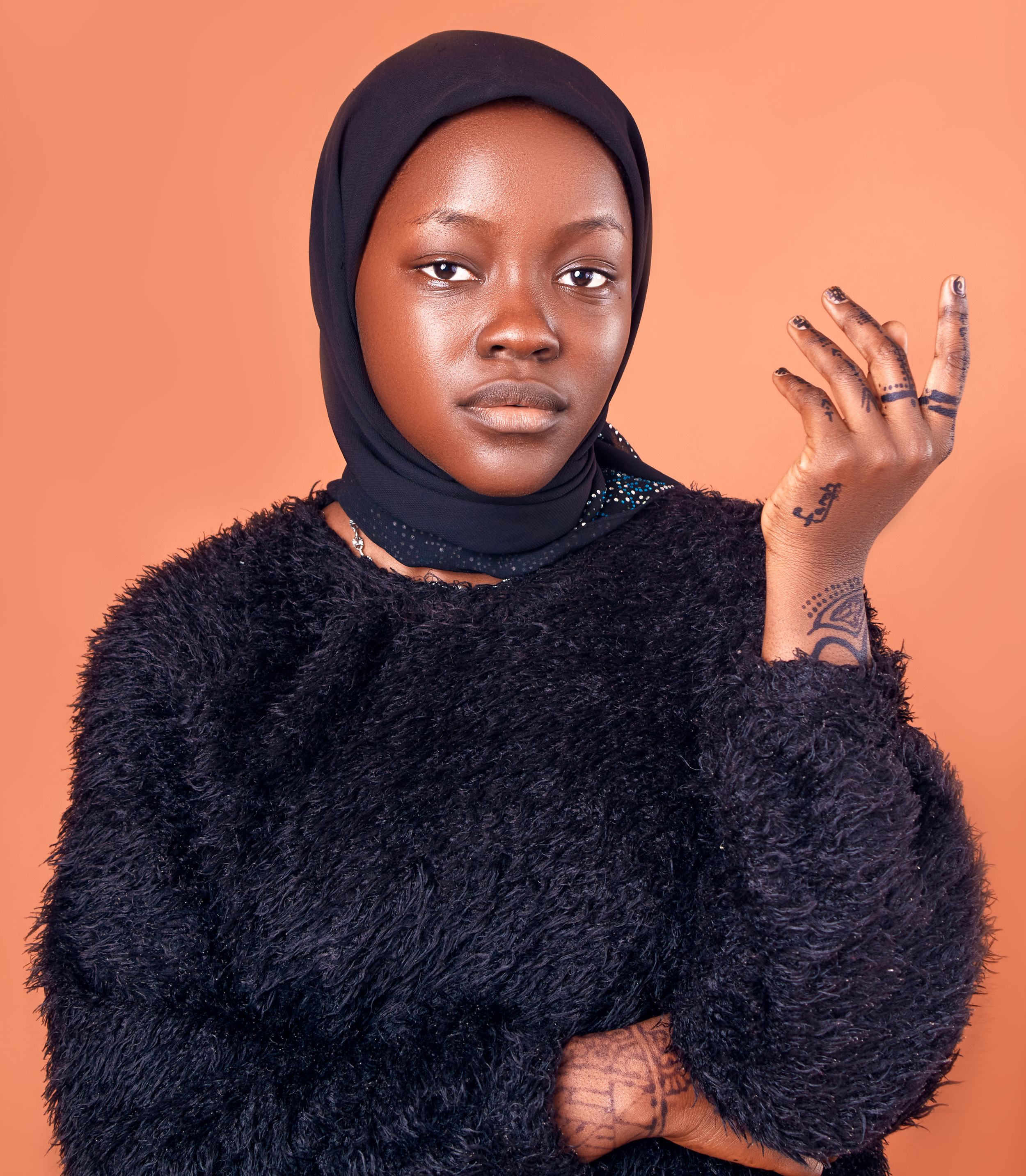 Stylish portrait of a woman in a black hijab and fluffy sweater against a peach background.