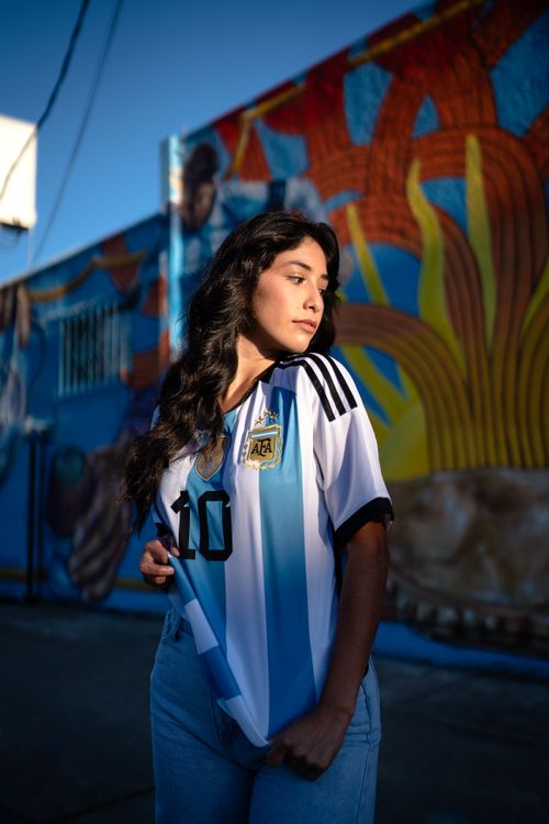 Free Portrait of a young woman wearing an Argentina football jersey, standing by a colorful mural during the day. Stock Photo