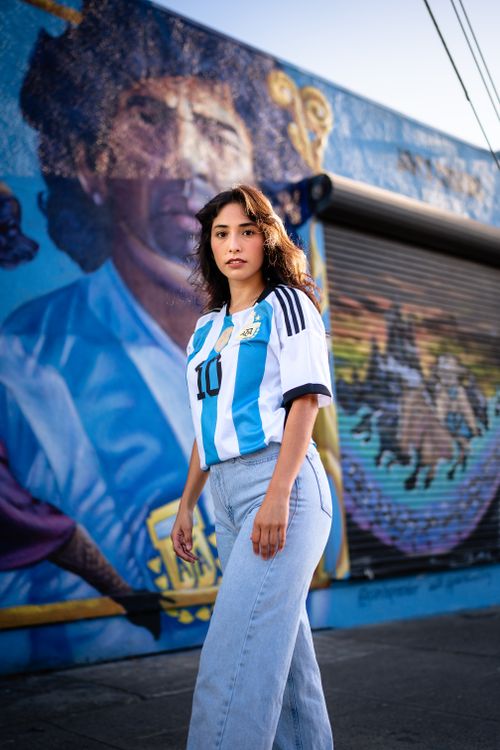 Free Confident young woman in an Argentinian football jersey poses in front of a vivid mural on a sunny city street. Stock Photo