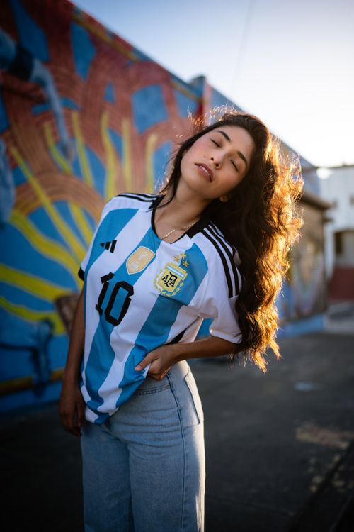Free Confident woman wearing a football jersey stands against a graffiti wall, sunlit alley view. Stock Photo