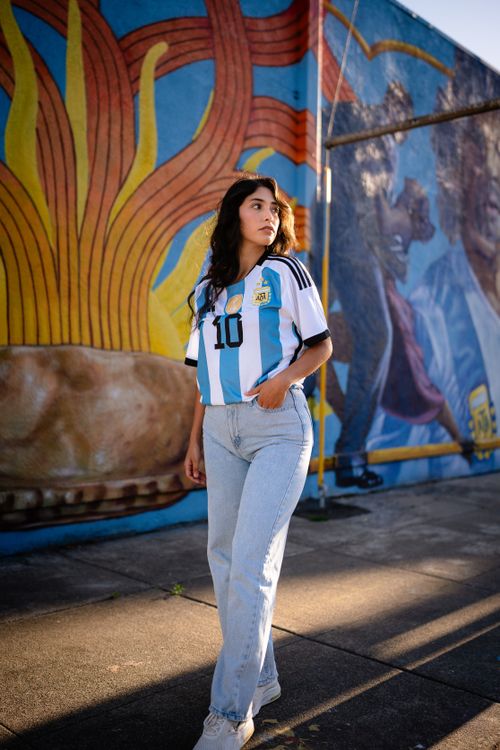 Free A woman stands in sunlight wearing an Argentina soccer jersey in front of a colorful mural. Stock Photo