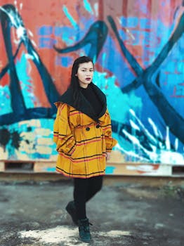 Confident woman in yellow coat poses against vibrant graffiti wall.