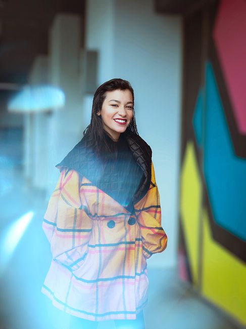 Stylish woman in vibrant coat posing in front of a colorful graffiti wall, smiling confidently.