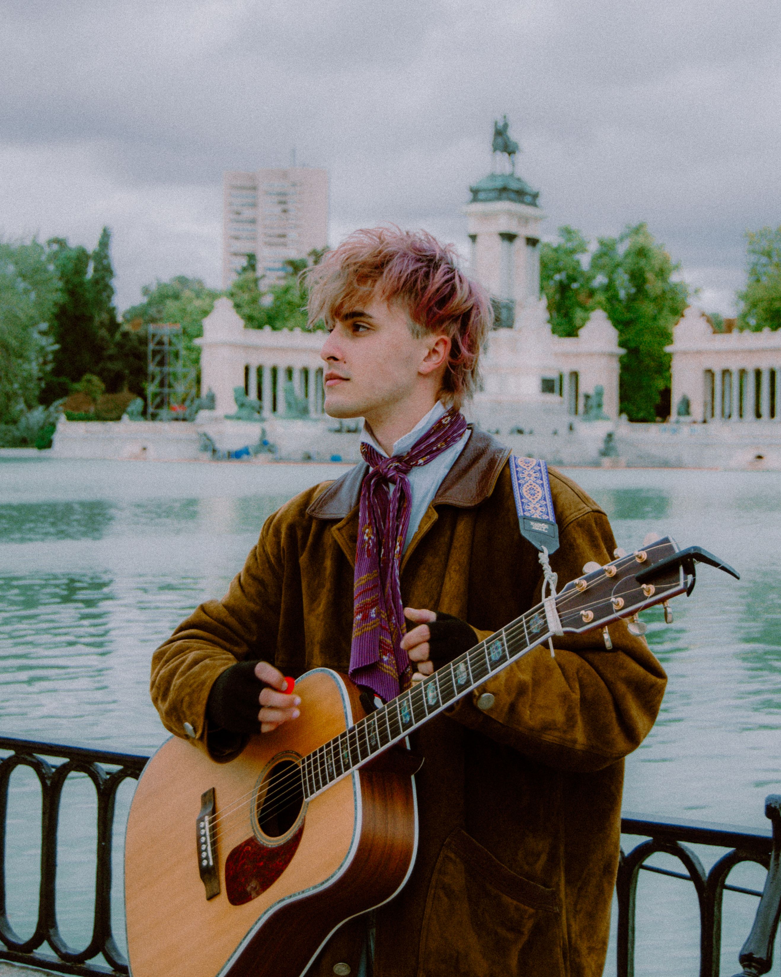 A young musician playing acoustic guitar by a scenic urban backdrop, offering artistic vibes.