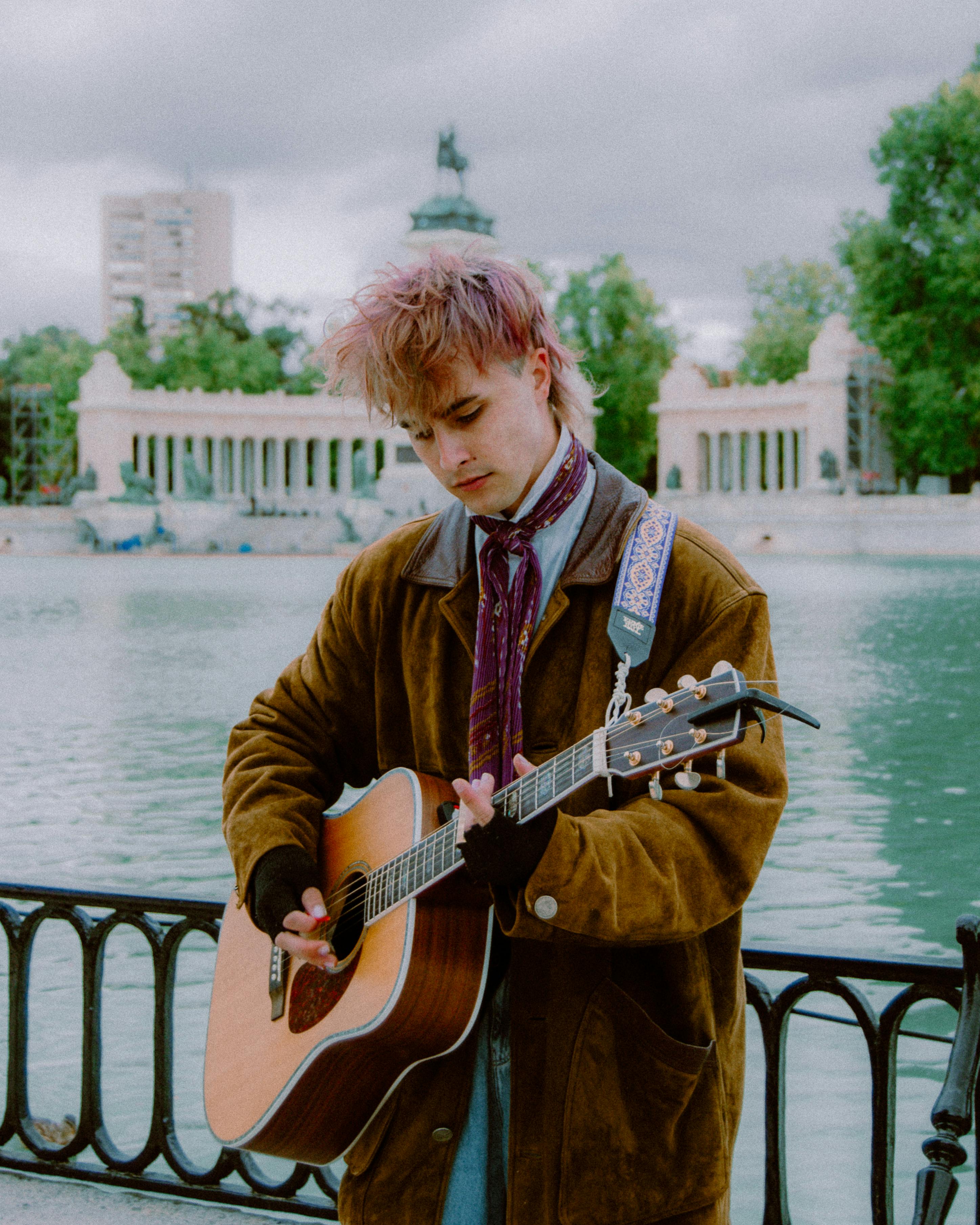 A young musician plays the acoustic guitar by a lake in a scenic park setting, creating a serene musical ambiance.