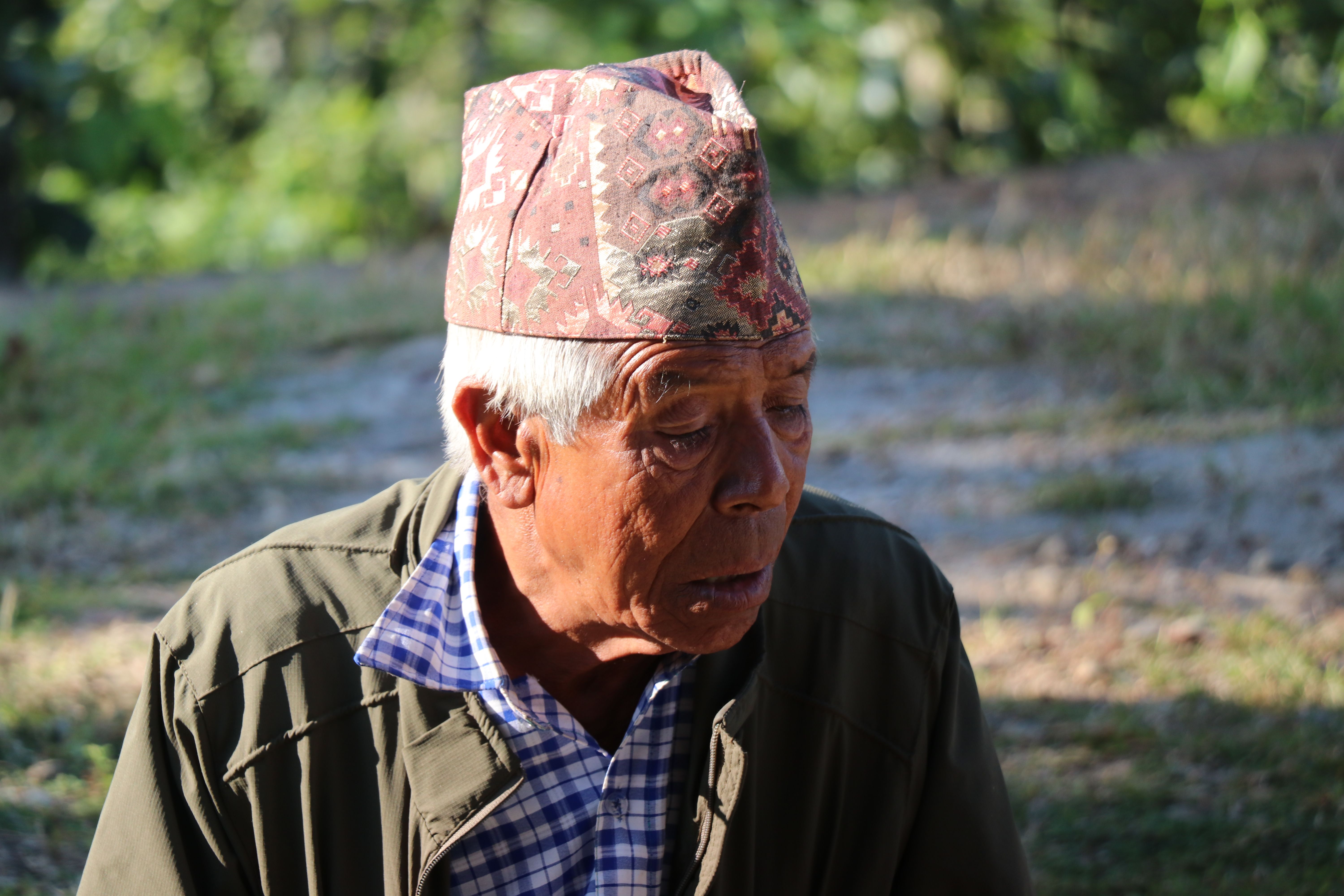 A senior man sitting outdoors wearing a traditional Nepali hat and jacket in a peaceful setting.