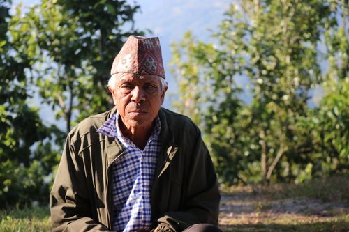 Free Elderly man wearing a traditional hat sitting outdoors with trees in the background. Stock Photo