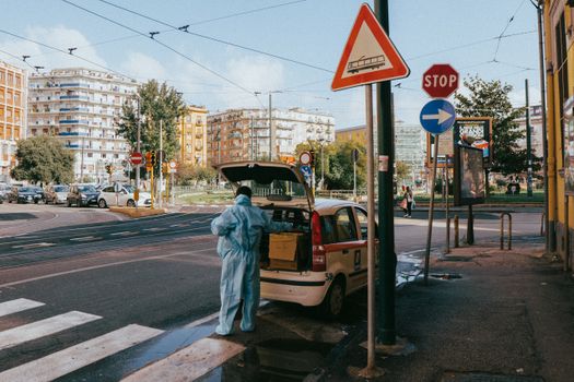 A healthcare worker in protective gear unloads a car in Neapolis, Italy, highlighting urban health efforts.