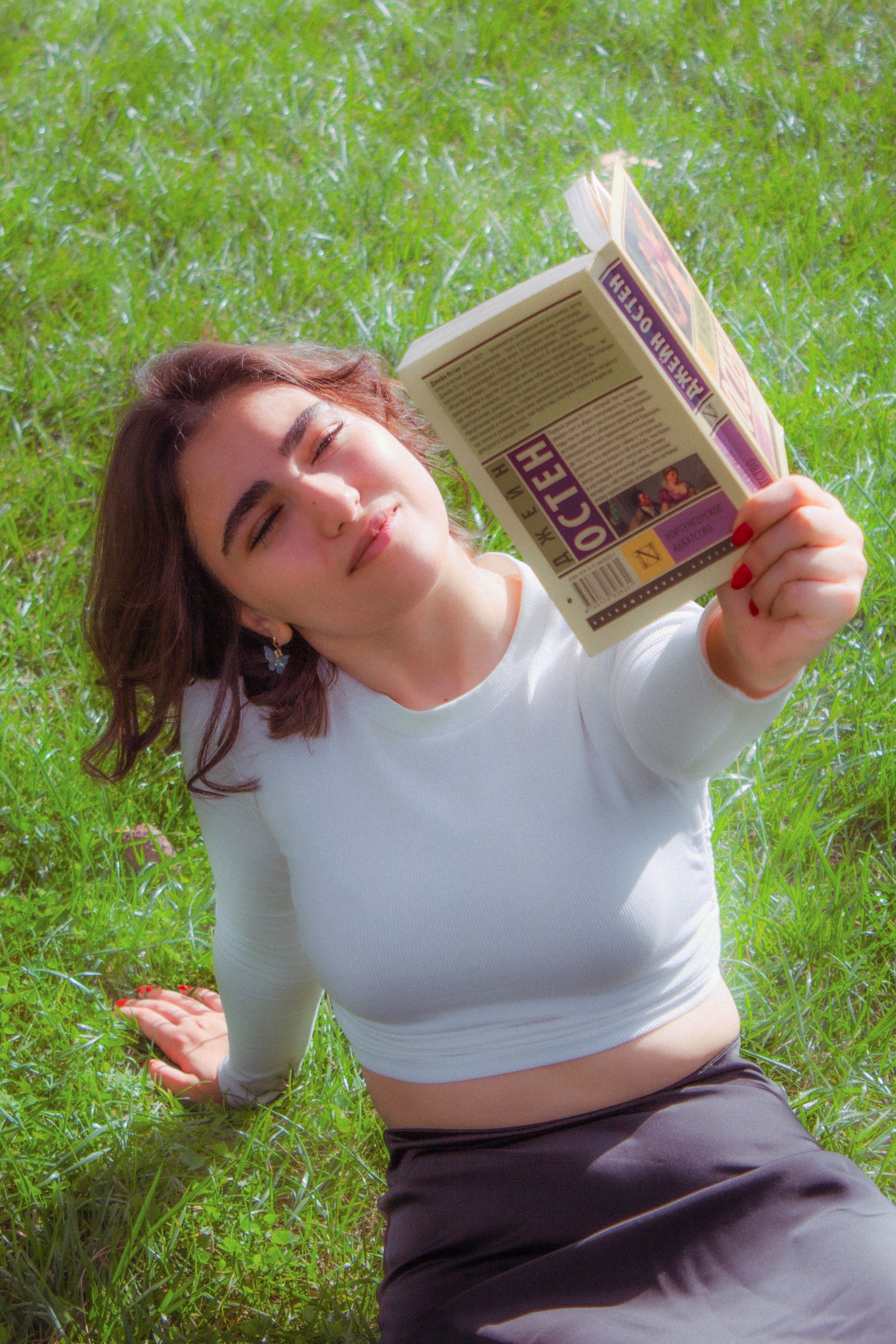 A woman in white reading a book on grass, captured on a sunny day.