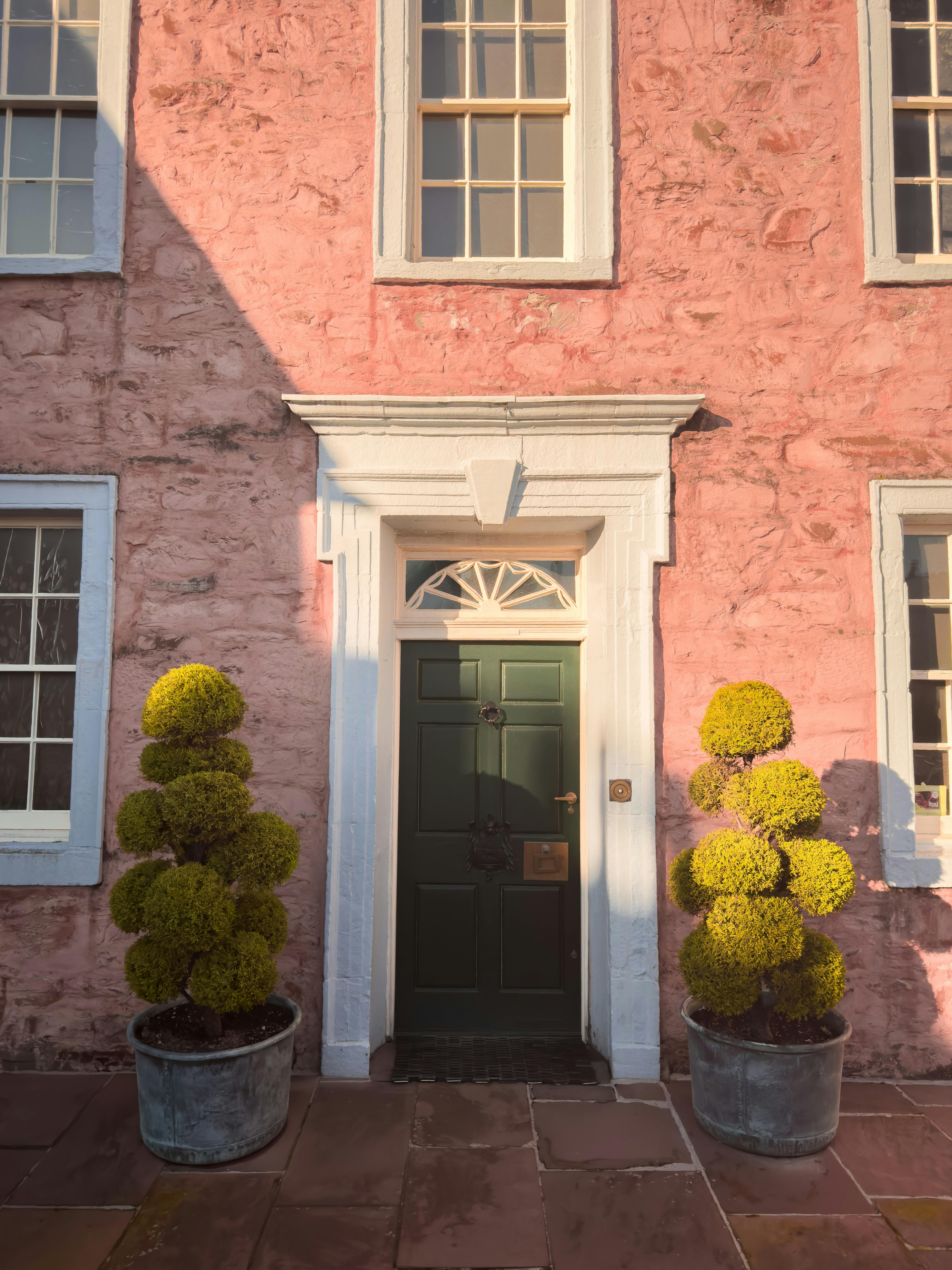 Pink townhouse facade with green door and potted plants flanking the entrance in warm sunlight.