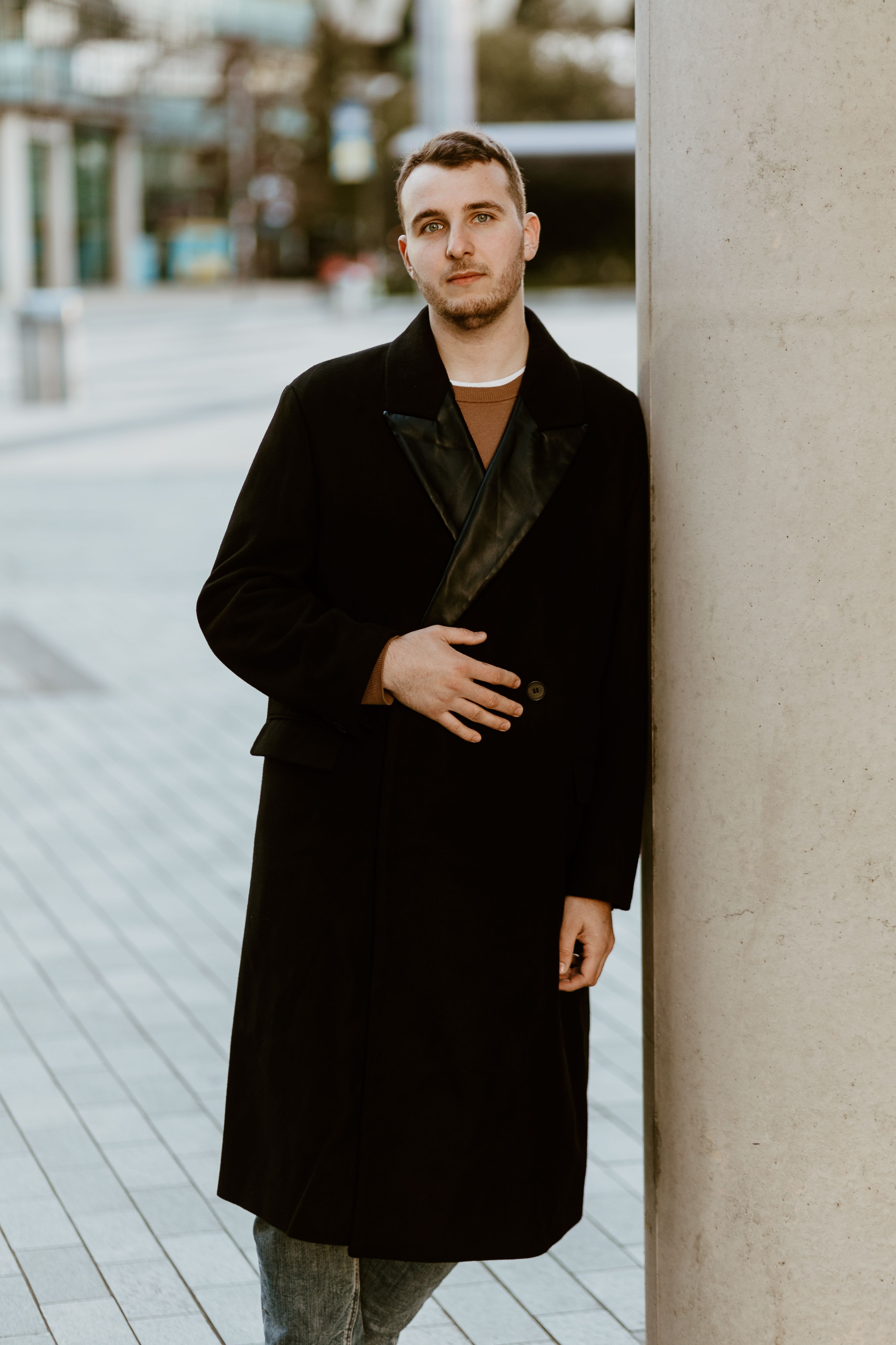 Stylish man in a long coat standing outdoors against a concrete pillar.