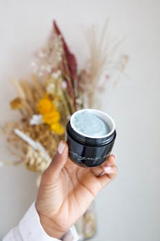Close-up of a hand holding a skincare cream jar with dried flowers in the background.