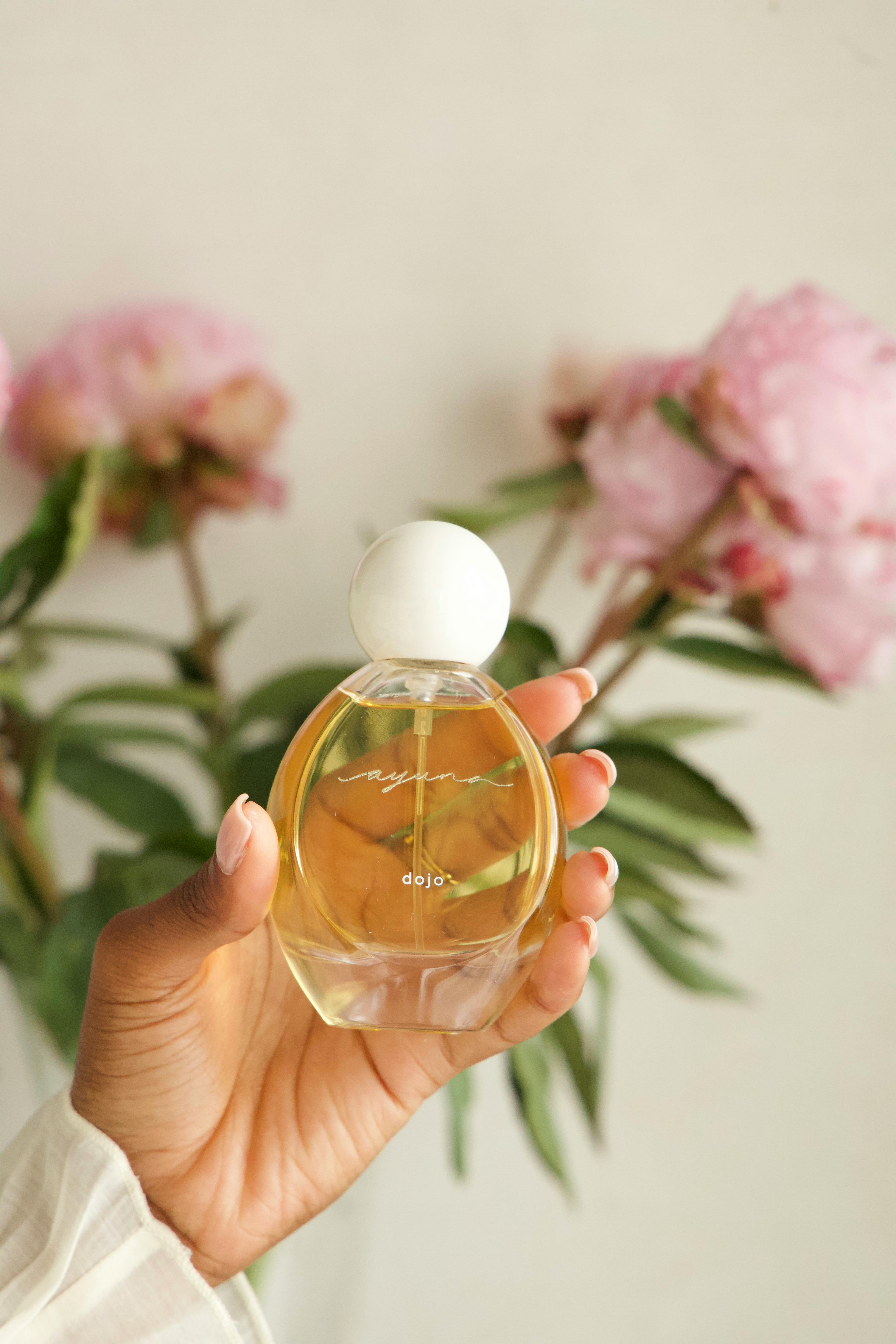 A hand holds a delicate perfume bottle with pink peonies in the background.
