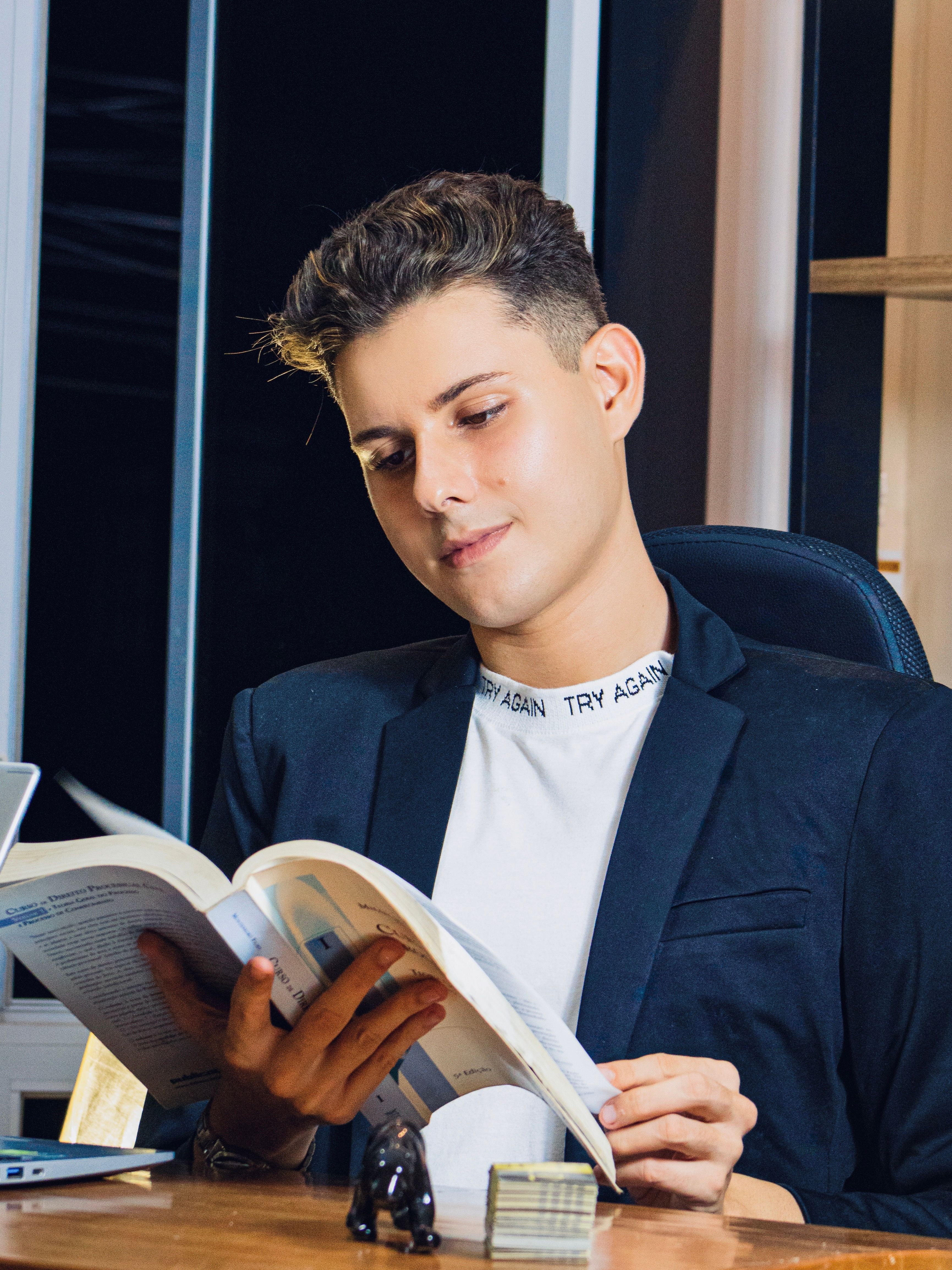 A young man in a blazer reads a book while sitting at a desk indoors. Modern setting.