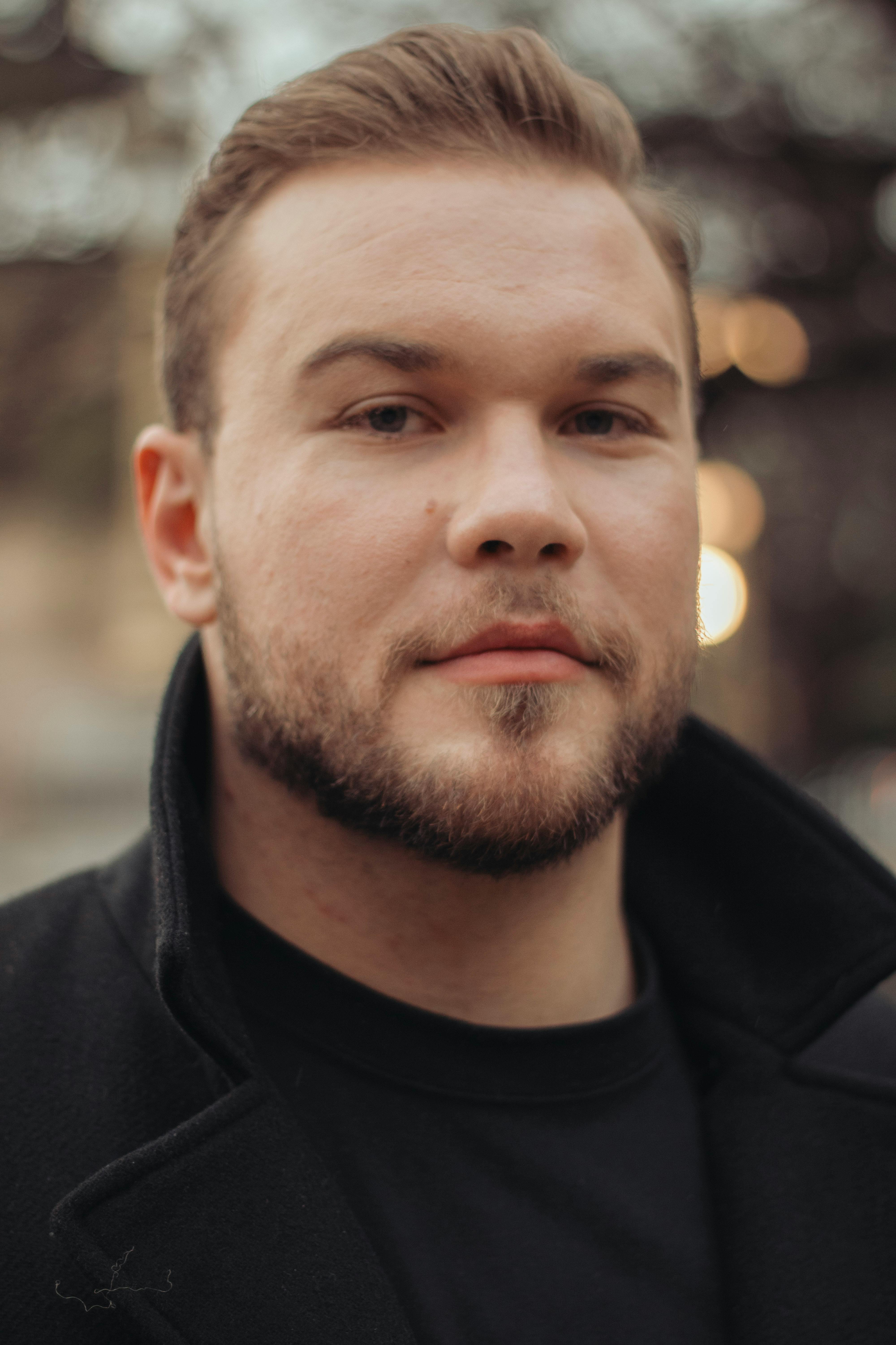 A handsome man with a beard in a black coat poses outdoors with a bokeh background.