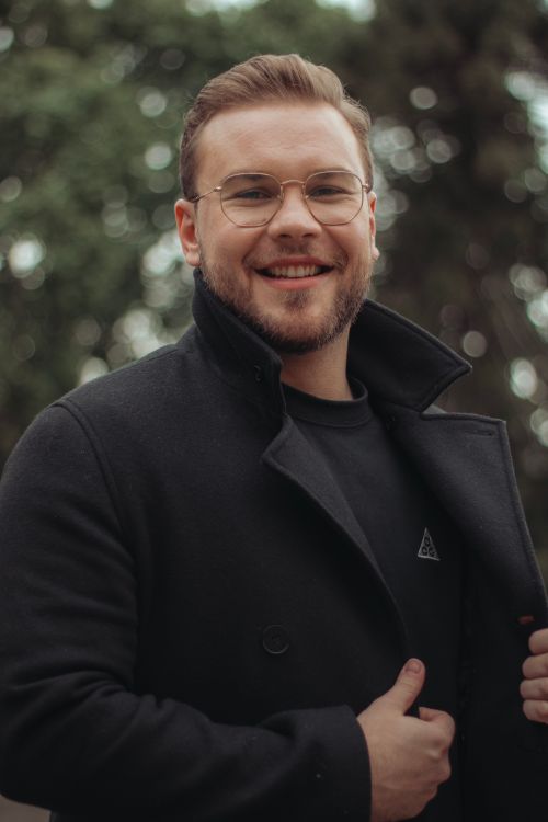 Free Cheerful man in glasses and black coat smiling outdoors on a cloudy day. Stock Photo