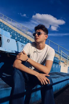 Young man in sunglasses and white t-shirt sits casually in blue stadium seats under clear sky.