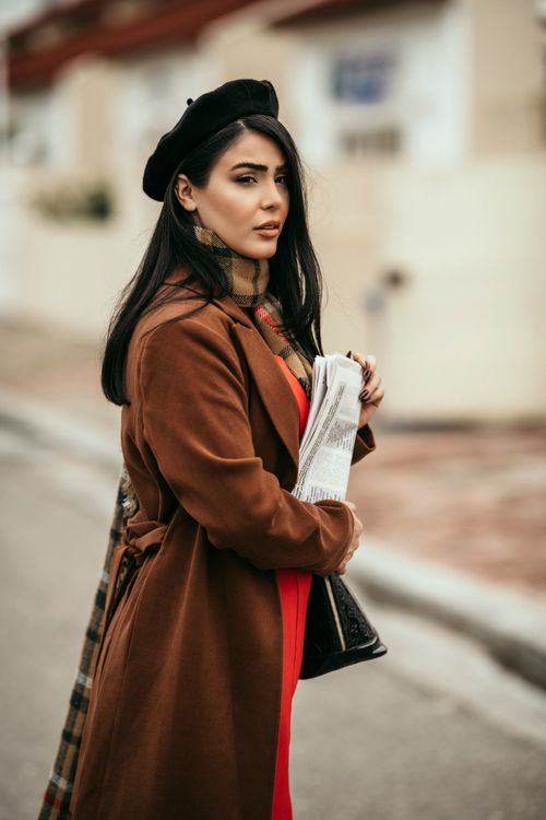 Free Stylish woman in a brown coat and beret holding a newspaper, standing in an urban setting. Stock Photo