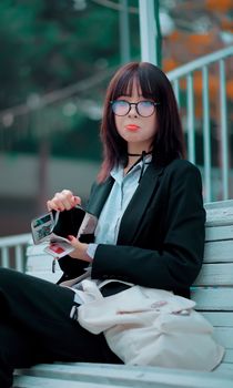 Stylish woman in glasses sitting on a bench with a briefcase outdoors.