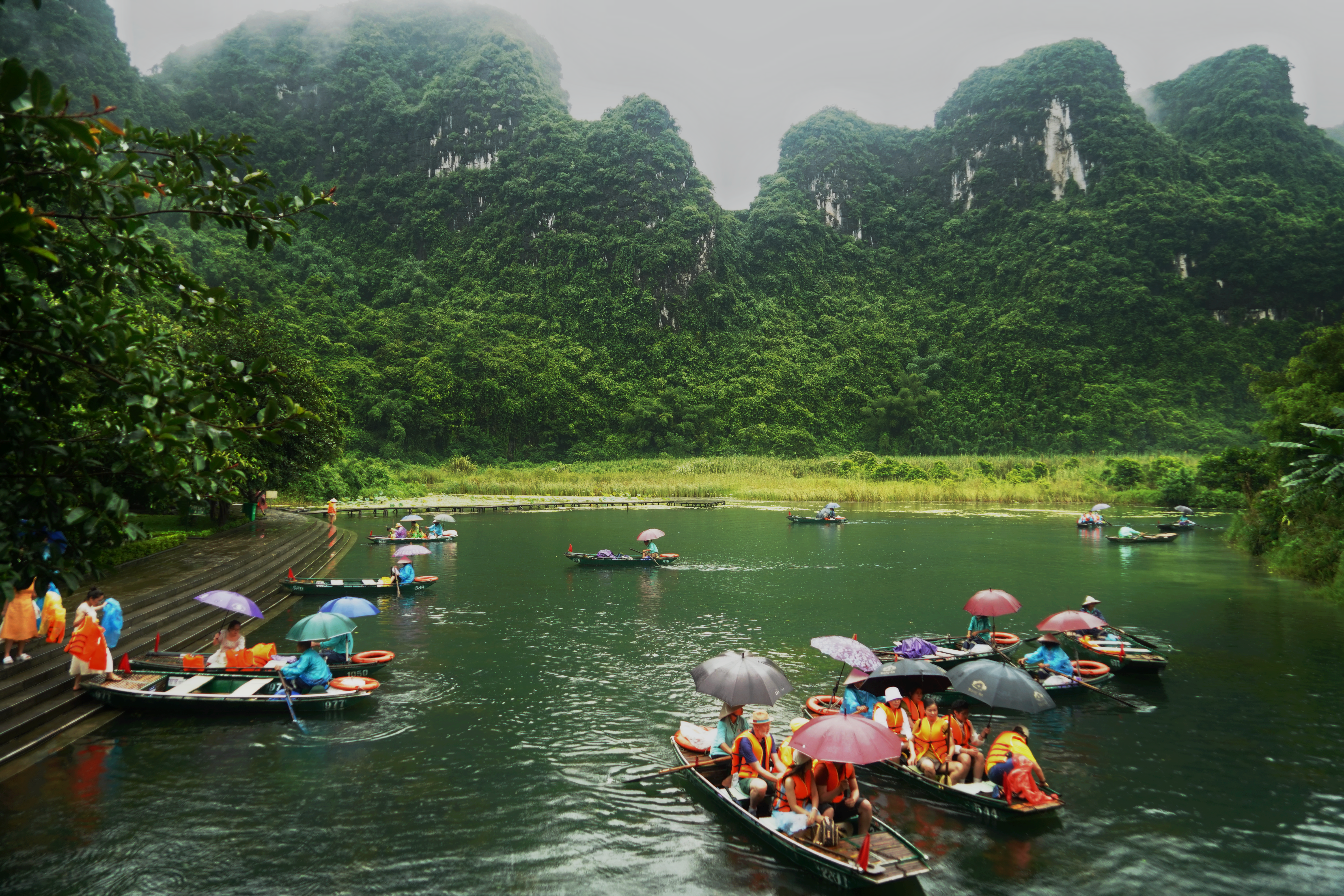 Groups of tourists in colorful boats on a scenic lake surrounded by lush greenery.