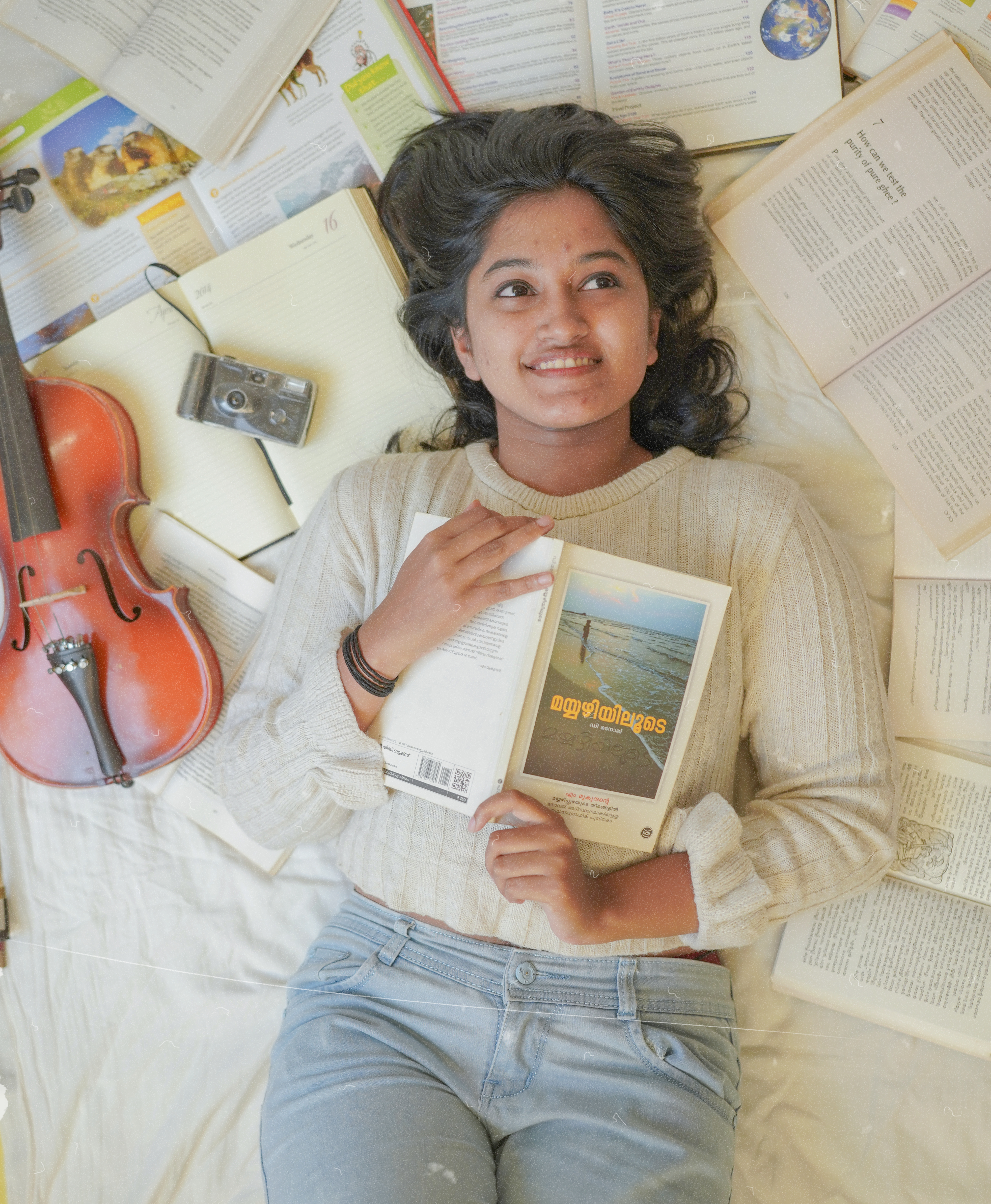 Smiling teenager lying on bed with book, violin, and camera surrounded by open books.
