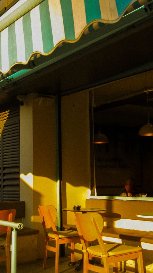 Free A cozy urban café with wooden chairs and a striped sunshade, basking in warm sunlight on a city sidewalk. Stock Photo